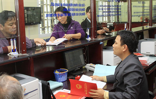 2 khách hàng đang đứng đợi làm thủ tục giấy tờ, một nhân viên ngồi trong quầy giao dịch đang cầm sổ đỏ